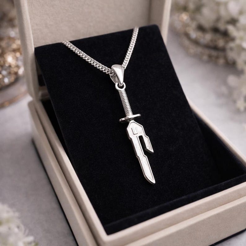 May include: A silver sword pendant necklace displayed in a gift box. The pendant features a detailed sword design with a textured handle and a stylized blade. The necklace chain is a delicate curb style.