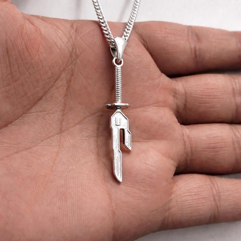 May include: A silver pendant necklace featuring a sword-shaped charm. The charm has a detailed hilt and blade design, suspended from a delicate chain. The necklace is shown against a neutral background.