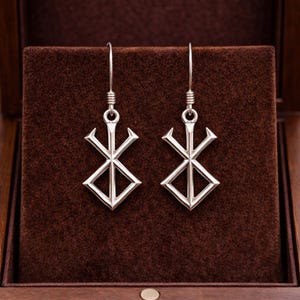 May include: A pair of silver-colored earrings featuring a geometric design. Each earring has a long hook and a stylized symbol with a cross-like shape above a diamond. The earrings are displayed in a brown velvet-lined box.