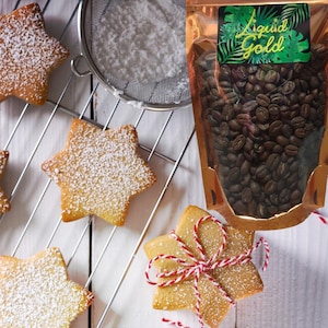Może przedstawiać: Ciasteczka maślane w kształcie gwiazdy posypane cukrem pudrem na metalowej kratce. Widoczna jest torebka z ziarnami kawy z napisem "Liquid Gold". Słowo "Shortbread" jest napisane na dole obrazu.