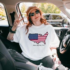 Puede incluir: Camiseta blanca con un mapa de los Estados Unidos con el diseño de la bandera americana. El texto "Since July 04, 1776" está impreso debajo. La camiseta la lleva una persona en un coche.