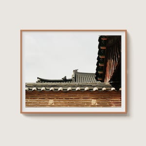 May include: Framed photograph of a traditional Asian building. The image features a section of the roof with dark gray tiles, decorative elements, and a textured, brown brick wall. The sky is a pale white.