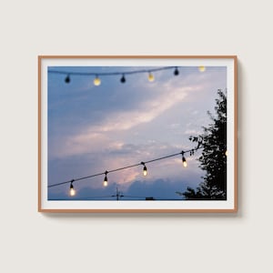 Può includere: Fotografia incorniciata di un cielo crepuscolare con luci a corda. Il cielo ha tonalità blu e rosa, con nuvole soffici. Una fila di luci rotonde illuminate è appesa attraverso la cornice. Rami di alberi in silhouette sono visibili a destra.
