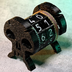 May include: A black and gold skull-shaped combination lock with a rotating dial featuring white numbers. The lock is secured with a gold-colored chain. The lock is resting on a gray surface.