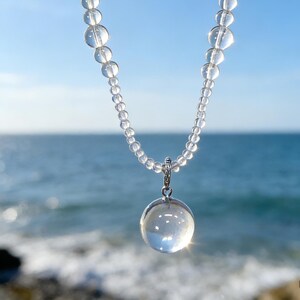 May include: A clear quartz crystal necklace with a large, round pendant. The necklace features small, clear beads strung together, with a larger, spherical pendant hanging from the center. The background is a bright blue sky and ocean.