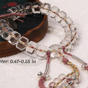 Puede incluir: Pulsera de cristal de cuarzo transparente con cuentas en forma de cubo, que mide 1,2-1,4 cm de diámetro. La pulsera presenta cordones rosas y multicolores, detalles metálicos y un dije decorativo. Las cuentas están ensartadas en un cordón transparente.