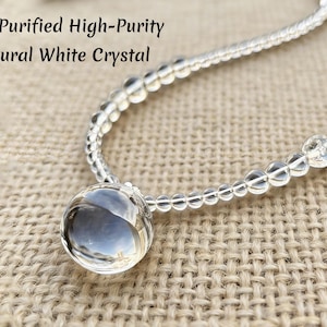 May include: A clear, natural white crystal necklace with a large, round pendant. The necklace is made of small, clear beads and is displayed on a textured, tan surface. Text on the image reads: "Fully Purified High-Purity Natural White Crystal."