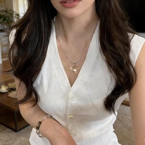 May include: A white sleeveless button-up vest is worn with a silver necklace and a bracelet. The necklace has a small, clear pendant. The bracelet features clear and black beads. The NineX Terra logo is in the bottom right corner.