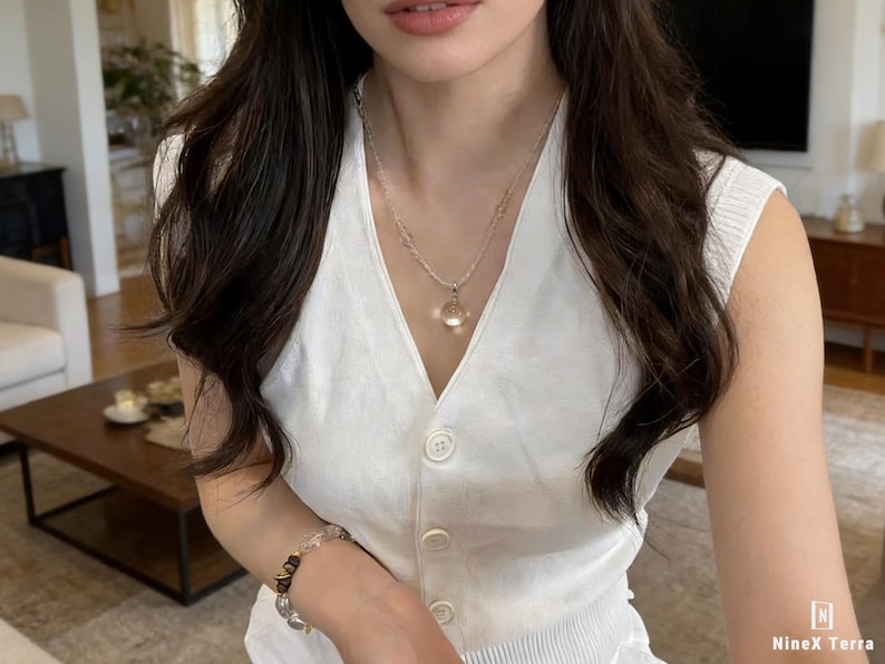 May include: A white sleeveless button-up vest is worn with a silver necklace and a bracelet. The necklace has a small, clear pendant. The bracelet features clear and black beads. The NineX Terra logo is in the bottom right corner.