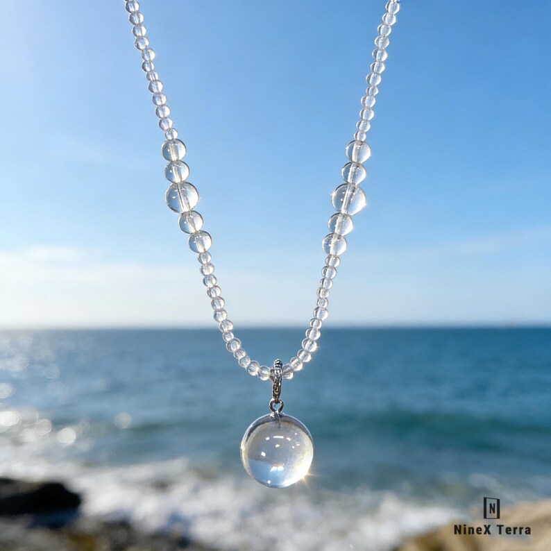 May include: A clear quartz crystal necklace with a large, round pendant. The necklace features small, clear beads strung together, with a larger, spherical pendant hanging from the center. The background is a bright blue sky and ocean.