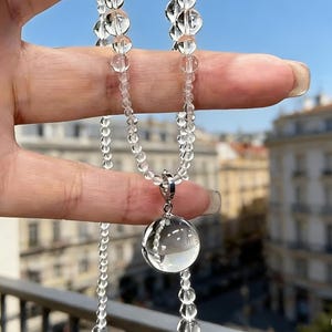 May include: A clear quartz crystal necklace with a large, round pendant and smaller beads. The necklace is held up against a blurred background of buildings and a blue sky. The pendant is approximately 1 inch in diameter.