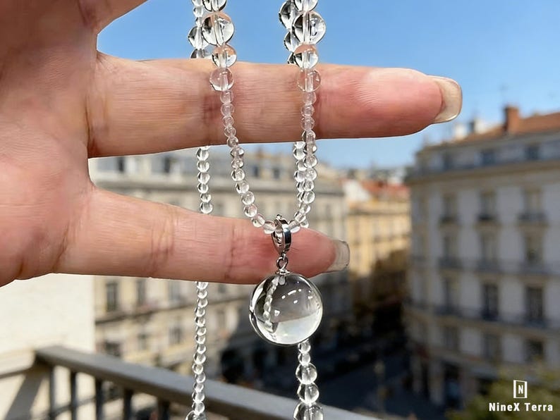 May include: A clear quartz crystal necklace with a large, round pendant and smaller beads. The necklace is held up against a blurred background of buildings and a blue sky. The pendant is approximately 1 inch in diameter.