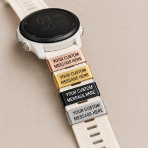 May include: A white Garmin smartwatch with a textured silicone strap featuring four rectangular metal charms in rose gold, gold, black, and silver. Each charm is engraved with the text YOUR CUSTOM MESSAGE HERE in a clean sans-serif font.