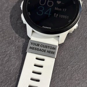 May include: A white Garmin smartwatch with a black bezel and a white band. The band features a silver plate with the text "YOUR CUSTOM MESSAGE HERE". The watch displays the time, date, and other metrics.