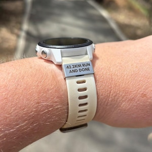 May include: A white and black smartwatch with a beige strap is shown on a wrist. The strap has the text "42.2KM RUN AND DONE" on a silver plate. The watch is designed for fitness tracking.