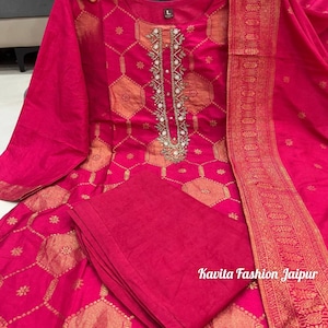 May include: A vibrant fuchsia kurta set featuring a gold hexagonal pattern and intricate silver embroidery along the neckline. The set includes a matching fuchsia dupatta with a wide gold border and solid fuchsia trousers. Text reads Kavita Fashion Jaipur.