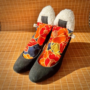 May include: A pair of black suede heels with a unique design. The interior features a vibrant floral pattern in red, orange, and yellow against a navy blue background. The top of each shoe is adorned with a white, textured material.