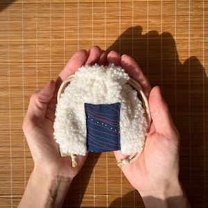 Może przedstawiać: Mała, puszysta biała torba ze sznurkiem i granatową prostokątną naszywką. Naszywka ma czerwony, zakrzywiony wzór linii i małe białe kropki. Torba jest trzymana w otwartych dłoniach.