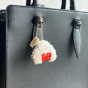 以下が含まれることがあります： 黒い質感のあるハンドバッグに、金色のチェーンで取り付けられた白くふわふわした三角形のおにぎり型のチャーム。チャームには小さな赤いアクセントがあり、側面には黒い文字でMUSUBIと印刷された白い布製のタグが付いています。
