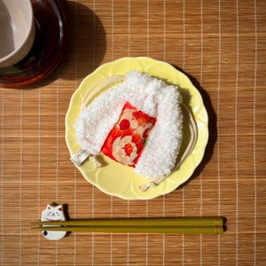 May include: A white, fluffy pouch with a red and floral-patterned insert rests on a yellow plate. The plate sits on a bamboo mat, with green chopsticks and a cat-shaped chopstick rest. A small tag with text is attached to the pouch.