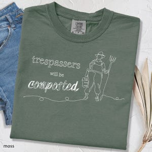 May include: Moss green t-shirt with the text "trespassers will be composted" in white script. A line drawing of a gardener holding a bucket and pitchfork is also featured on the shirt. The shirt is folded.