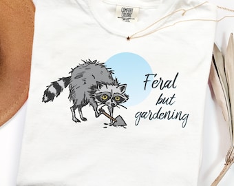 Può includere: T-shirt bianca con un procione grigio con una pala, su un cerchio azzurro. Il testo "Feral but gardening" è stampato sulla maglietta. È visibile anche una collana d'oro con un ciondolo a stella.