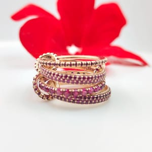 Peut inclure: Une pile de bagues dorées, chacune ornée d'une bande de petites pierres précieuses rouges rondes. Les bagues sont empilées les unes sur les autres. Une fleur rouge est en arrière-plan.