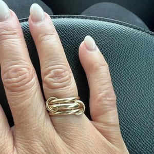 May include: A gold-colored ring with a unique design, featuring three interconnected bands. The ring is worn on a finger, showcasing its intricate details and polished finish. The ring's design is modern and eye-catching.