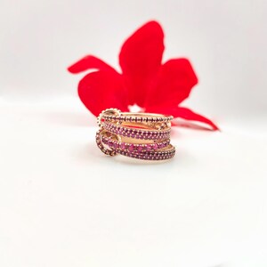 May include: A stack of gold-colored rings with multiple bands, each encrusted with small, vibrant red gemstones. The rings are set against a white background with a blurred red flower in the background, creating a striking contrast.