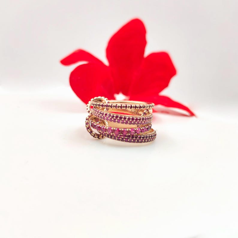 May include: A stack of gold-colored rings with multiple bands, each encrusted with small, vibrant red gemstones. The rings are set against a white background with a blurred red flower in the background, creating a striking contrast.