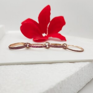 May include: Four gold-toned rings with small, red gemstones are linked together. The rings are arranged in a row on a white surface, with a bright red flower in the background. The rings are delicate and elegant.