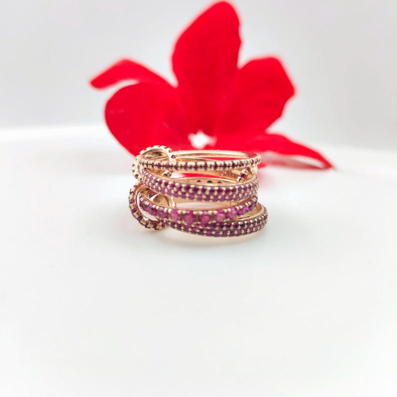 May include: A stack of gold-colored rings with a band of small, round, red gemstones. The rings are stacked on top of each other. A red flower is in the background.