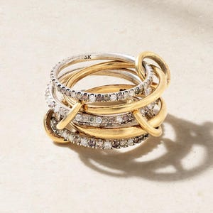 Peut inclure: Une bague multi-bandes avec des anneaux dorés et argentés entrelacés. Certains anneaux sont sertis de petites pierres scintillantes. La bague présente un design superposé unique, avec les initiales "SK". L'esthétique générale est élégante et sophistiquée.