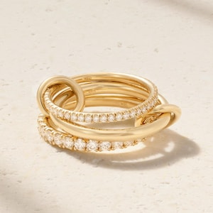 Puede incluir: Un conjunto de anillos dorados con un diseño entrelazado. Un anillo está adornado con pequeñas gemas brillantes. Los anillos son de color oro cálido y se exhiben sobre una superficie clara. Los anillos son una joya.