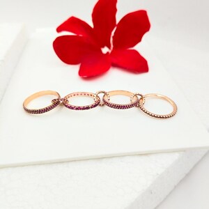 May include: A linked ring set with four rose gold rings. Three rings are adorned with small, red gemstones, while the fourth ring has a textured design. A red flower is in the background.