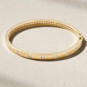 May include: A gold-colored bangle bracelet with square-cut, clear gemstones. The bracelet has a hinged clasp and a series of small square cutouts along the top edge. The bracelet is displayed on a light-colored surface.