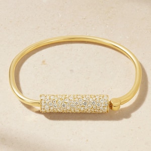May include: A gold-colored bangle bracelet with a cylindrical, diamond-studded center. The bracelet has a smooth, polished finish and a simple, elegant design. The diamonds sparkle, adding a touch of luxury to the jewelry.