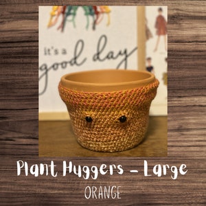 May include: Orange crocheted plant hugger with black button eyes. It's a good day text is visible in the background. Plant Huggers - Large ORANGE.