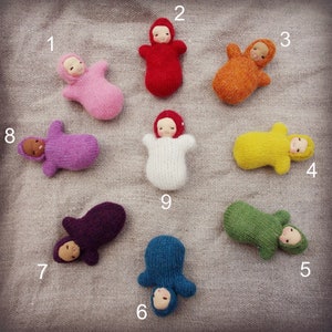 Può includere: Nove bambole di neonato in maglia fatte a mano in vari colori, tra cui rosso, arancione, giallo, verde, blu, viola, rosa e bianco. Le bambole sono disposte in cerchio su uno sfondo di tessuto beige.