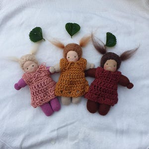 Może przedstawiać: Trzy ręcznie robione lalki z włosami z przędzy i szydełkowymi sukienkami w kolorze różowym, pomarańczowym i bordowym. Każda lalka ma prostą twarz i jest ułożona na białej powierzchni z zielonymi liśćmi.