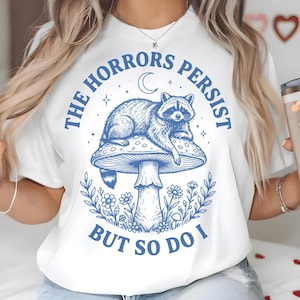 以下が含まれることがあります： キノコの上にいるアライグマの青いグラフィックが特徴の白いTシャツ。テキスト「THE HORRORS PERSIST BUT SO DO I」がプリントされています。ハートのステッカーが付いたステンレス製のタンブラーも表示されています。