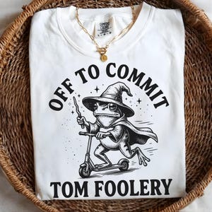 Könnte beinhalten: Weißes T-Shirt mit einem schwarz-weißen Aufdruck einer Frosch-Illustration mit Hexenhut und Umhang, der Roller fährt. Der Text lautet "OFF TO COMMIT TOM FOOLERY". Eine goldene Halskette liegt auf dem Shirt.