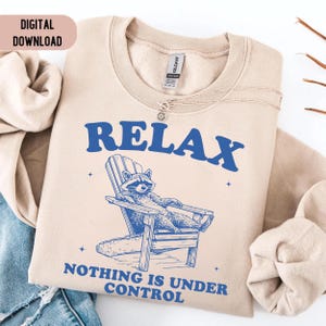 May include: Beige sweatshirt with the word "RELAX" in blue block letters. A raccoon is lounging in a blue Adirondack chair. The text "NOTHING IS UNDER CONTROL" is below the raccoon. A digital download tag is in the upper left corner.