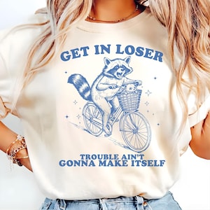 Könnte beinhalten: Cremefarbenes T-Shirt mit einer blauen Illustration eines Waschbären auf einem Fahrrad mit einem Baby-Waschbären in einem Korb. Der Text "GET IN LOSER" steht über dem Waschbären und "TROUBLE AIN'T GONNA MAKE ITSELF" darunter.