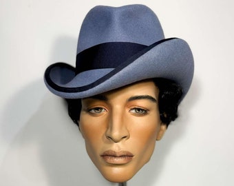 Puede incluir: Una cabeza de maniquí con un sombrero fedora azul y sosteniendo una cámara negra vintage con manos de madera.