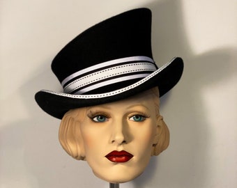 Puede incluir: Un sombrero de copa negro con una banda de rayas blancas y negras. El sombrero está inclinado hacia un lado y está sentado en una cabeza de maniquí.