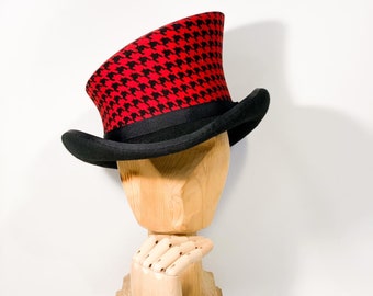 Puede incluir: Un sombrero de copa negro con un patrón de pata de gallo rojo y negro en la corona. El sombrero está inclinado hacia un lado y está sentado en un soporte de sombrero de madera.