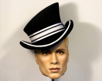 Puede incluir: Un sombrero de copa negro con una banda blanca y costuras blancas. El sombrero está inclinado hacia un lado y está en una cabeza de maniquí.