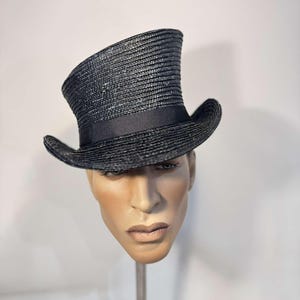 May include: A black top hat with a slightly angled crown and a black ribbon band. The hat is made of woven material and has a classic, formal design. The hat is displayed on a mannequin head.