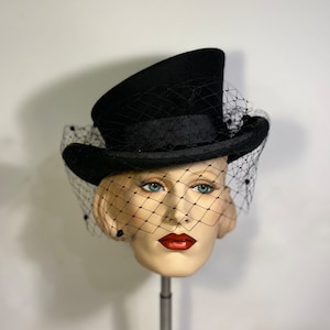 May include: A black top hat with a black veil. The hat is angled to the side and the veil is draped over the hat.